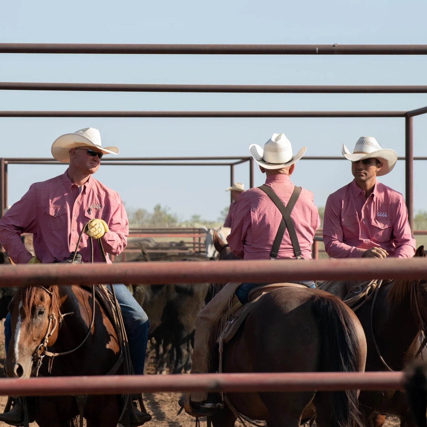 Three cowboys on horseback in a pen wearing pink shirts and white hats.