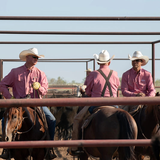 Three cowboys on horseback in a pen wearing pink shirts and white hats.