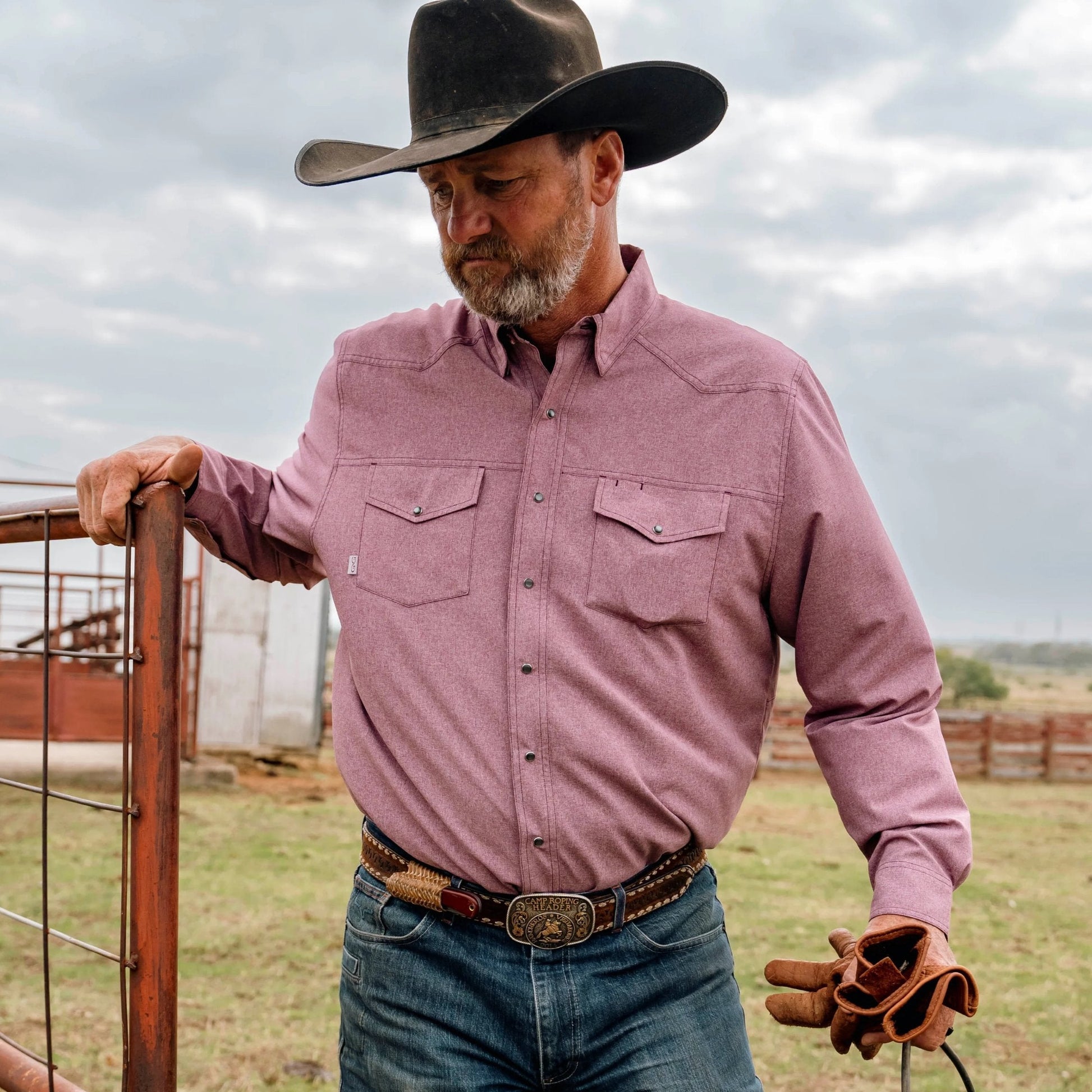 Man in cowboy attire standing in a field with a cloudy sky.
