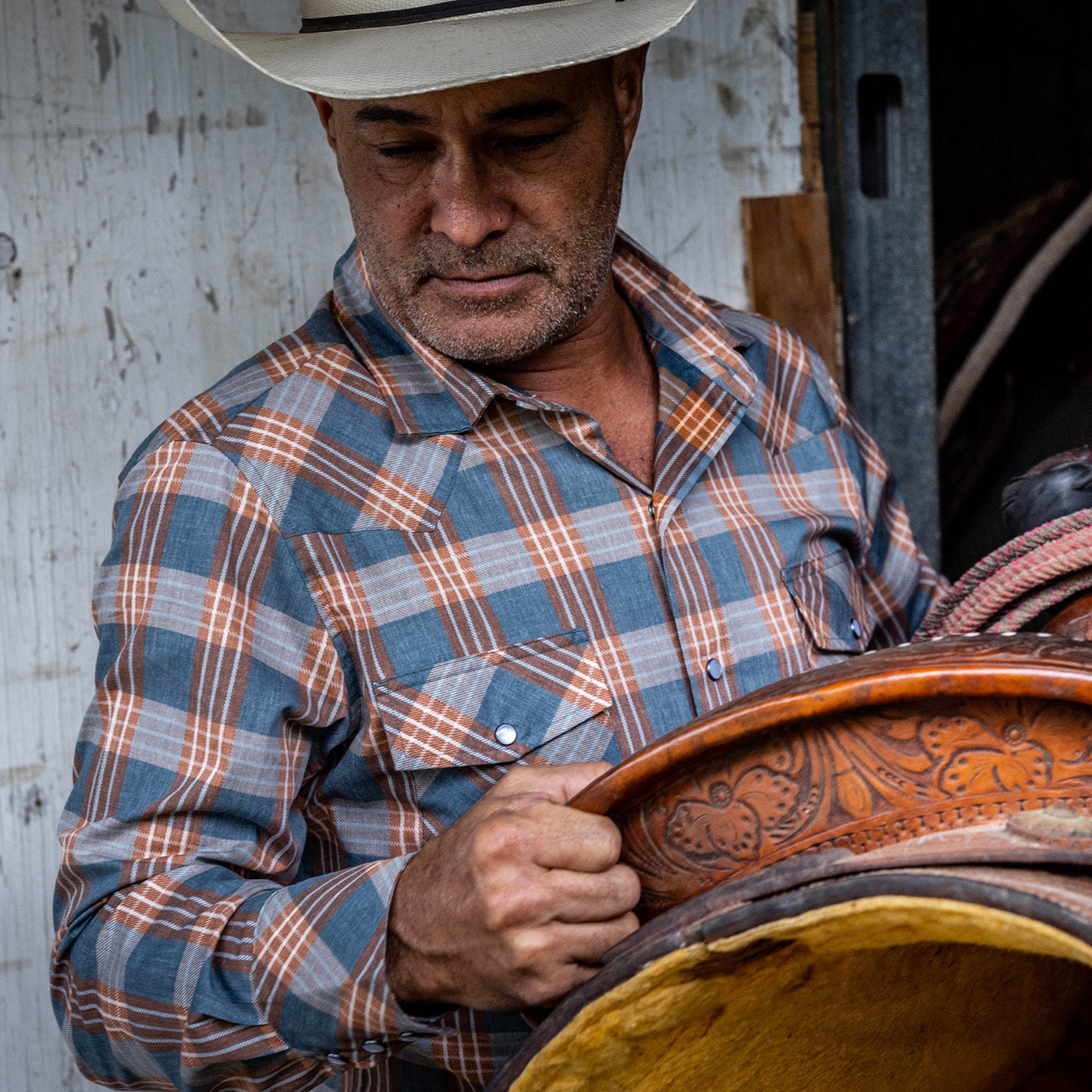Man in plaid shirt and cowboy hat holding a leather saddle