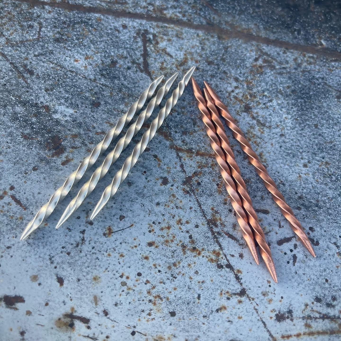 Two sets of twisted metal toothpicks, one silver and one copper, on a textured concrete surface.
