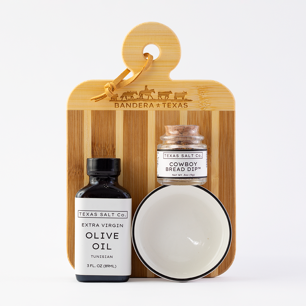 Texas Salt Co. olive oil bottle, cowboy bread dip jar, and ceramic bowl on a wooden board with 'Banderas Texas' branding.